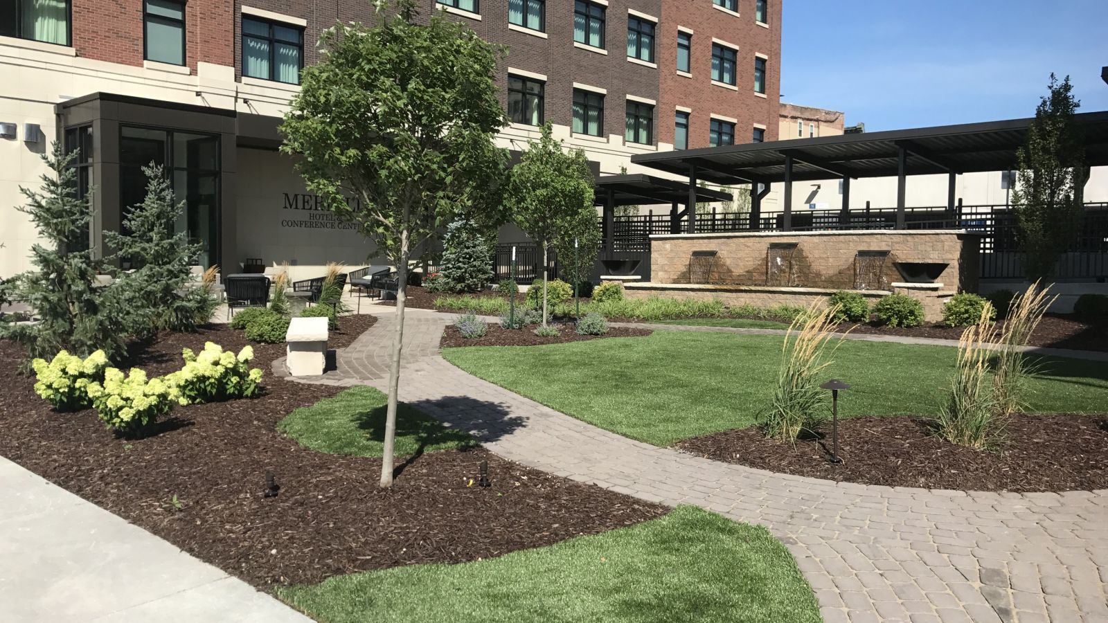 Brick pathway winds through a landscaped garden with trees, shrubs, and a water feature, adjacent to a multi-story building.