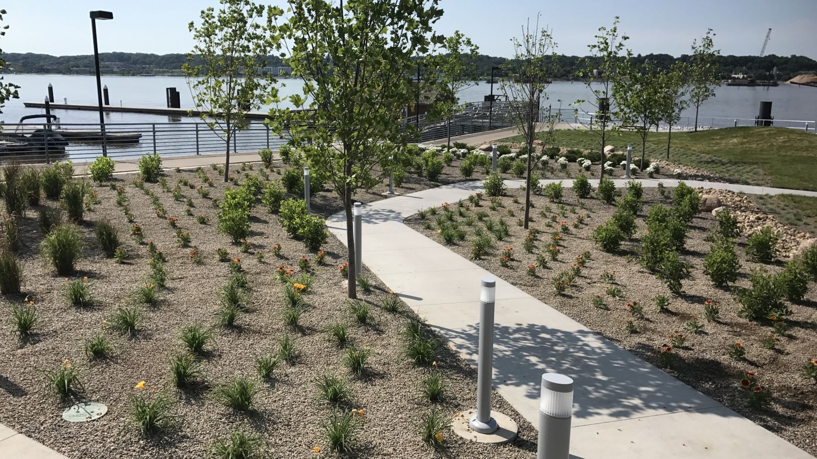 A paved walkway curves past landscaped beds of plants toward a lake with a dock.
