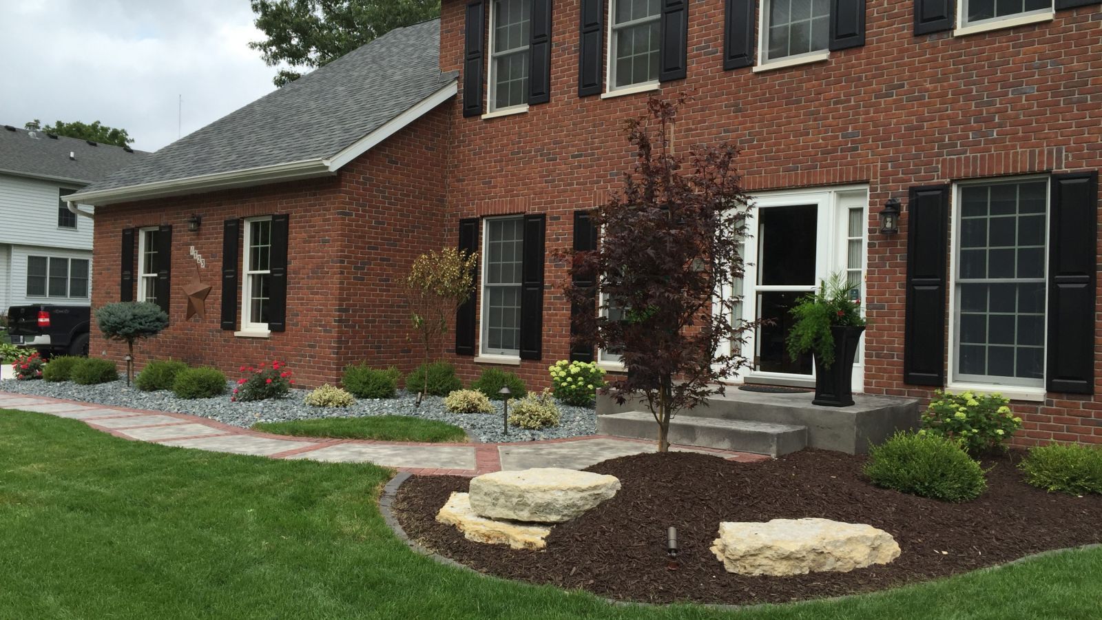 Brick house with black shutters and a landscaped front yard featuring bushes, trees, and a stone pathway with gray gravel.