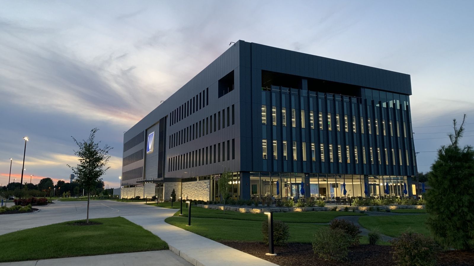 Modern, dark-gray office building with glass windows and a grassy lawn, illuminated at dusk.