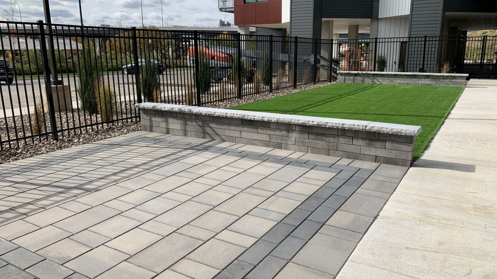 A paved patio area with a raised section of artificial turf, bordered by a retaining wall and a black fence.