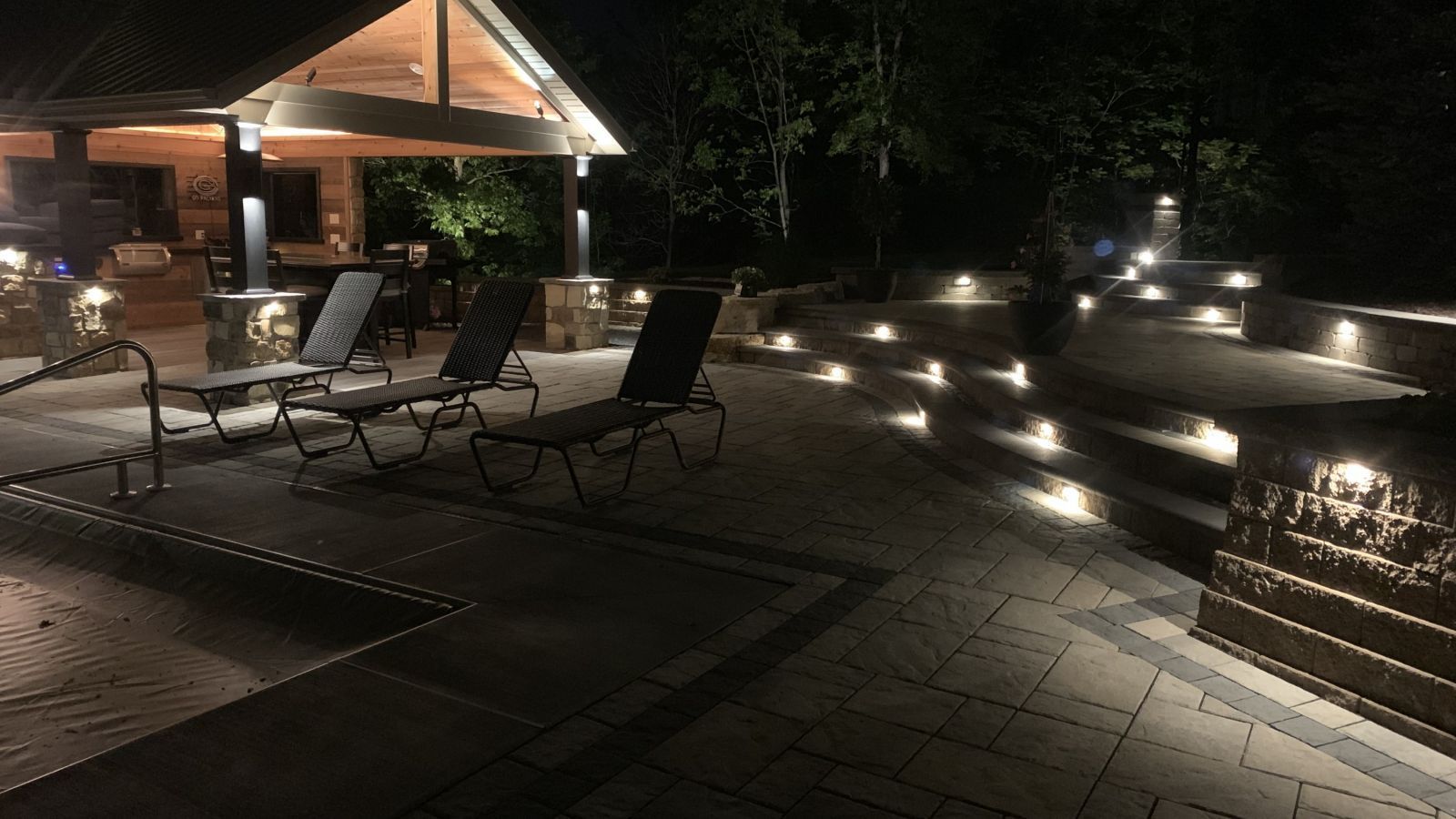 Nighttime outdoor patio with chaise lounge chairs and landscape lighting near a covered structure.