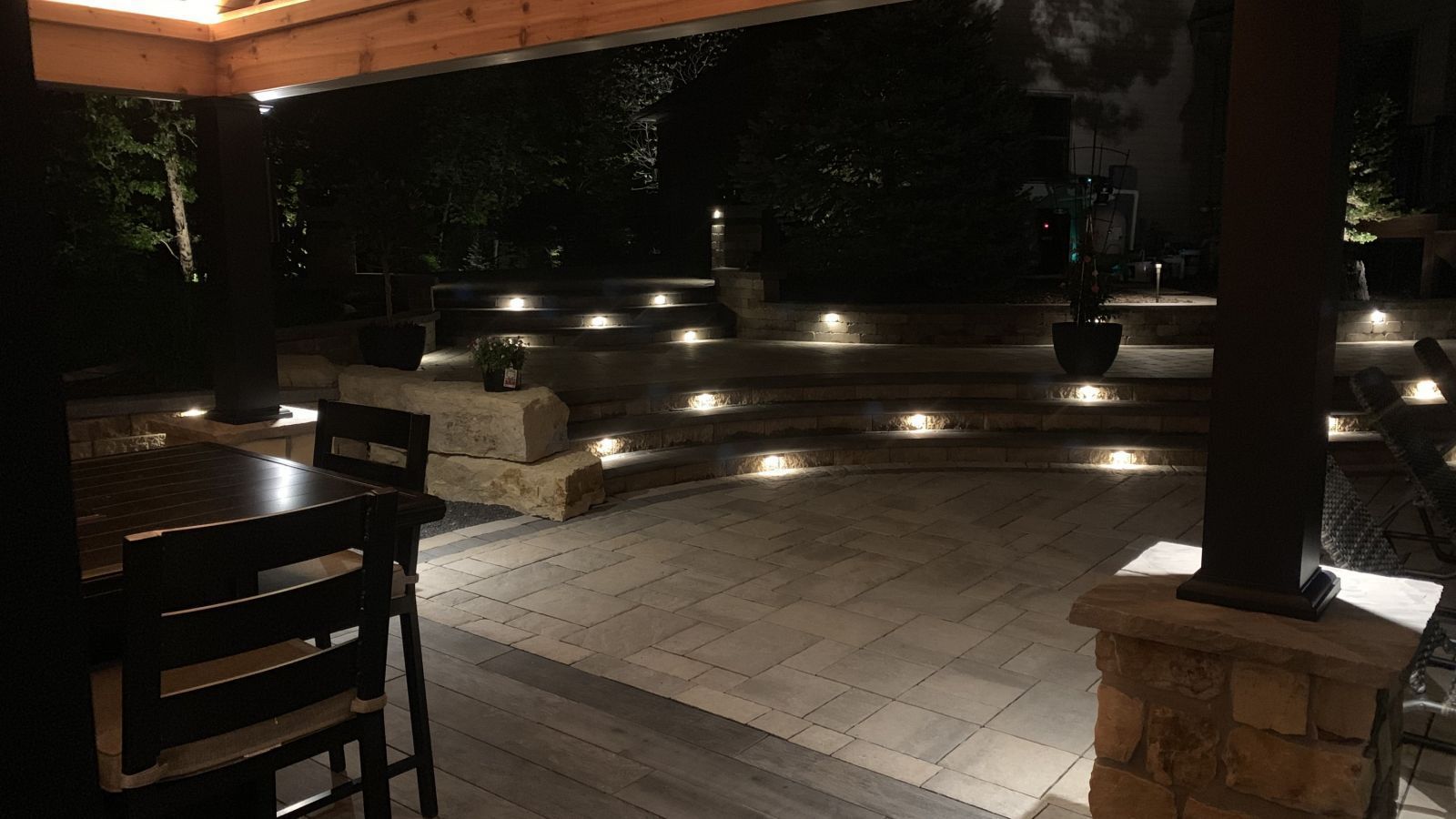 Nighttime outdoor patio with warm lighting illuminating steps and pillars, with a table and chairs in the foreground.