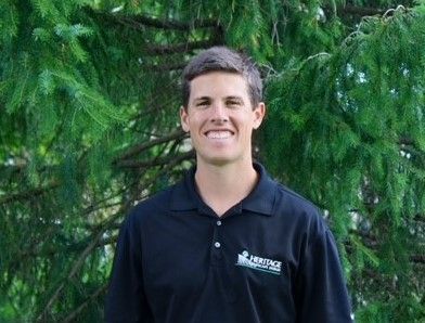 A man in a black polo shirt is smiling in front of a tree.