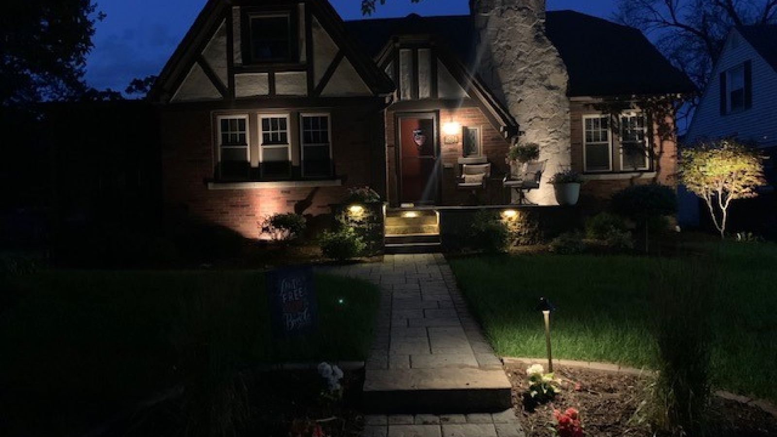 A Tudor-style house illuminated at night with landscaping lights along a brick pathway leading to the front door.