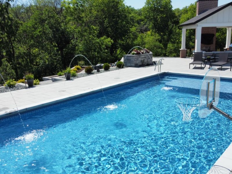 A rectangular swimming pool with fountains and a basketball hoop, surrounded by a patio and lush greenery.