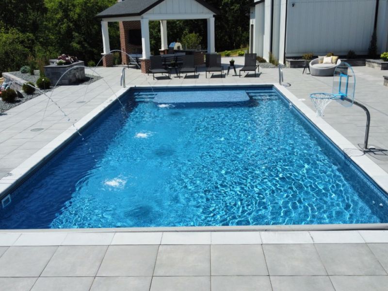 Rectangular outdoor swimming pool with blue water, a small hot tub area, and basketball hoop on a stone patio, with a gazebo and lounge chairs visible.