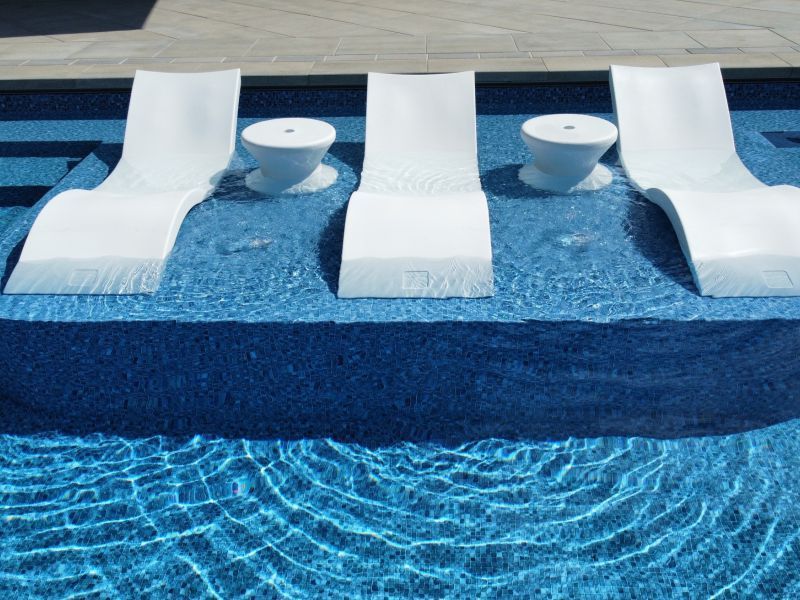 White chaise lounge chairs and side tables in a blue tiled pool. Water ripples in the foreground.