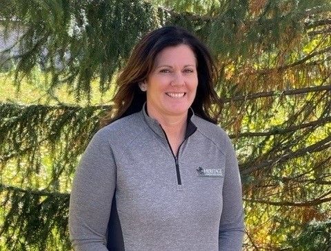 A woman in a gray shirt is smiling in front of a tree.