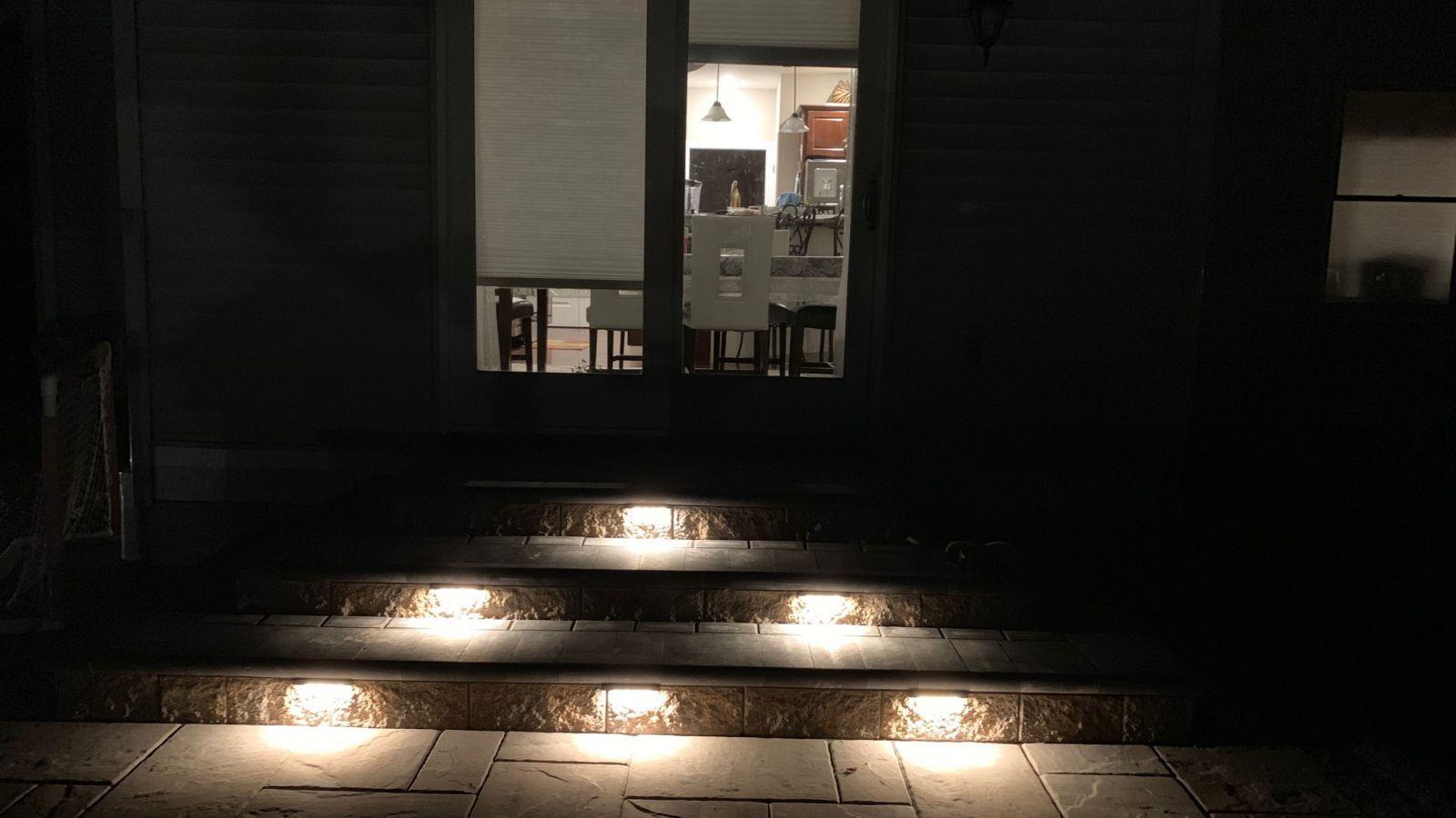 Lit steps leading up to a glass door at night. Interior light illuminates kitchen and dining furniture.