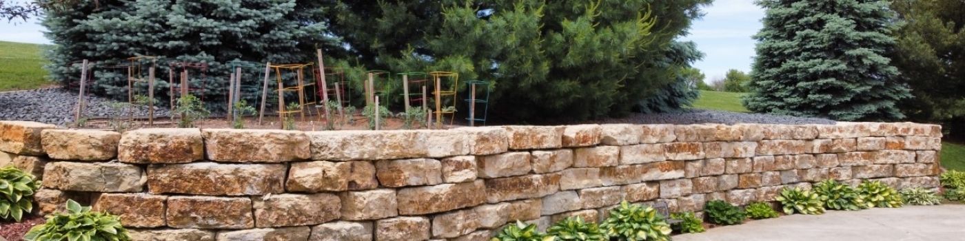 A long, tan stone wall borders a garden with greenery and trees in the background.