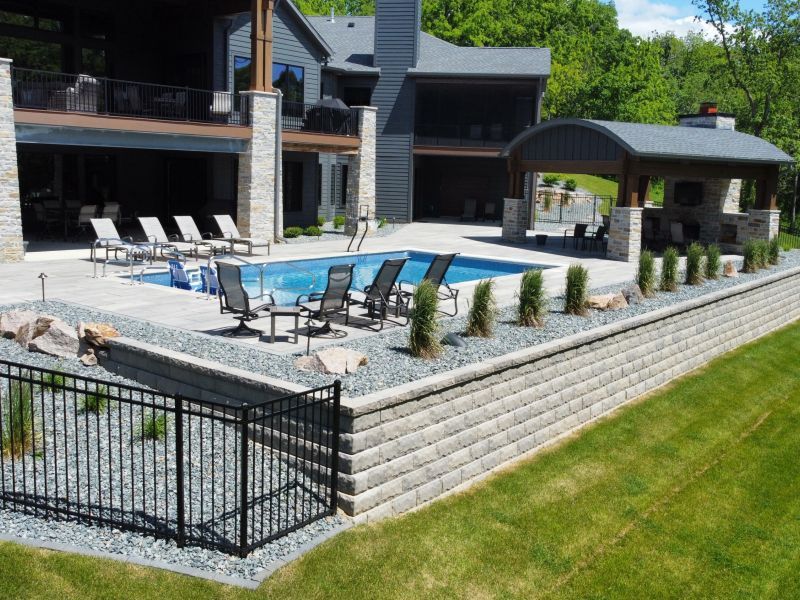 A luxurious backyard with a pool, patio, and outdoor kitchen set against a multi-story home with a green landscape.