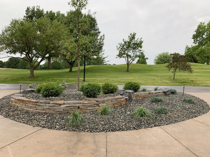 Circular landscaped garden bed with stone wall and gravel, surrounded by a paved path, with trees and grassy hill in the background on an overcast day.