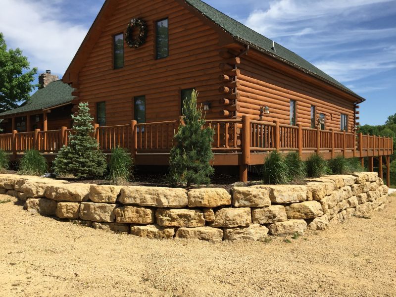 Log cabin with a stone retaining wall in front and a wooden deck. Sunny day.