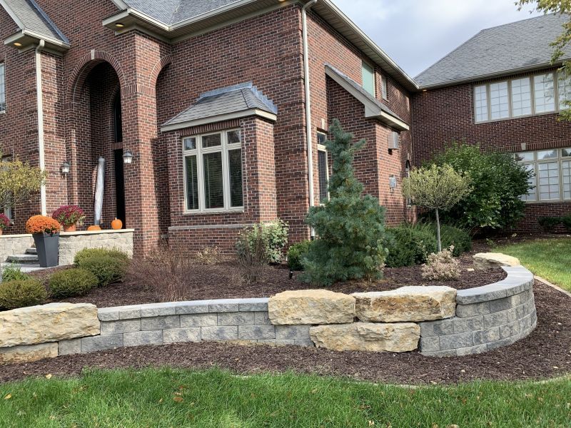 A brick house with a stone retaining wall in front of a landscaped yard. There are various plants and mulch beds.