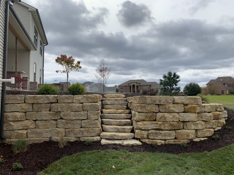 Stone retaining wall with steps leading up to a higher level, located near a house under a cloudy sky.