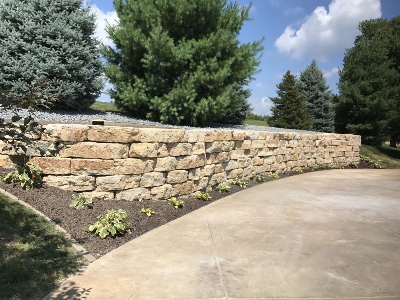 Stone retaining wall with planted flowers curving alongside a concrete driveway, with trees in the background under a blue sky.