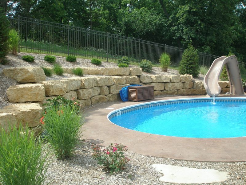 Pool with a curved slide, bordered by a stone retaining wall with landscaping and a sloped lawn in the background.