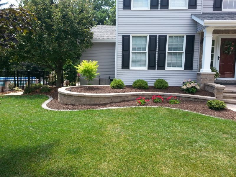 Front yard with a curved stone flower bed filled with mulch and flowers, in front of a gray house with black shutters.