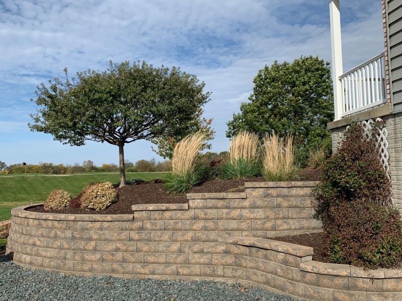 Brick retaining wall with tiered landscaping, trees, and ornamental grasses against a blue sky.