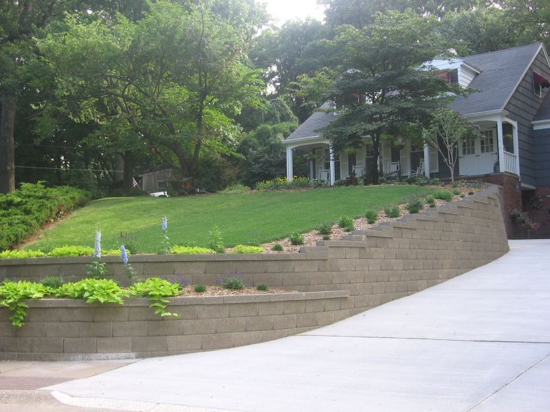 A tiered retaining wall next to a driveway leads to a house on a grassy hill with trees in the background.