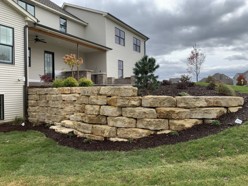 Stone retaining wall in front of a two-story house with a covered porch, built on a small grassy hill.