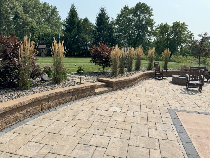 Stone patio with retaining wall, featuring ornamental grasses and seating area overlooking a lawn and trees on a sunny day.