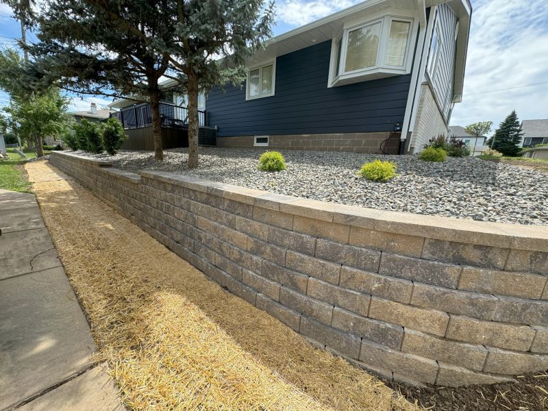 A house with a retaining wall along the front yard. The wall is made of tan bricks. The yard has gravel, grass and trees.
