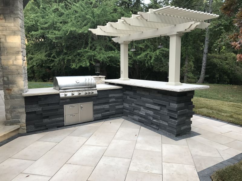 Outdoor kitchen with a grill and counter, gray stone facing, and a white pergola. Set on stone pavers with green trees in the background.