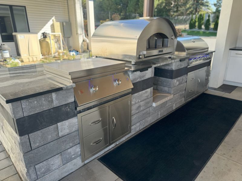 Outdoor kitchen with stainless steel grill, pizza oven, and stone countertops. Gray and black color scheme, set on a patio.