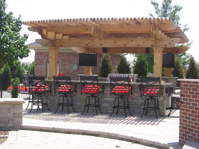 Outdoor bar with a stone facade, wooden pergola, and swivel bar stools with red cushions.