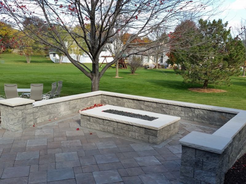 A stone patio with a built-in fire pit and surrounding walls. The backyard has green grass and trees.