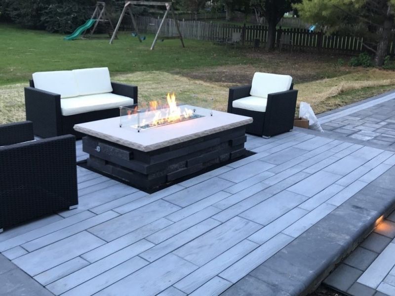 Outdoor patio with a fire pit and seating, including a love seat and two chairs, on a paved patio with a grassy background.