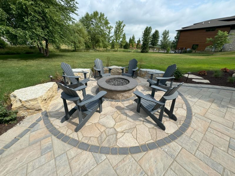 A circular fire pit with gray chairs arranged around it, set on a stone patio in a backyard. Lush green lawn and trees in the background.