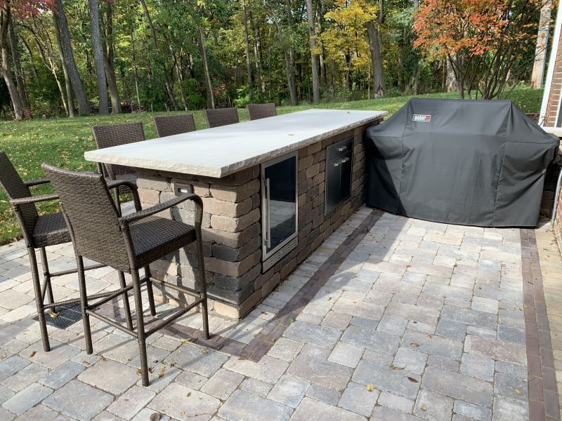Outdoor kitchen with a stone island, concrete countertop, and seating. A covered grill sits nearby.