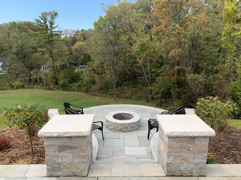 A patio with a fire pit and seating surrounded by landscaping, overlooking a grassy yard and trees.