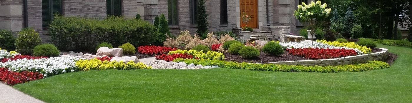 A vibrant flower bed in front of a building, featuring a mix of red, yellow, white, and green plants. Lush green lawn surrounds the flowerbed.