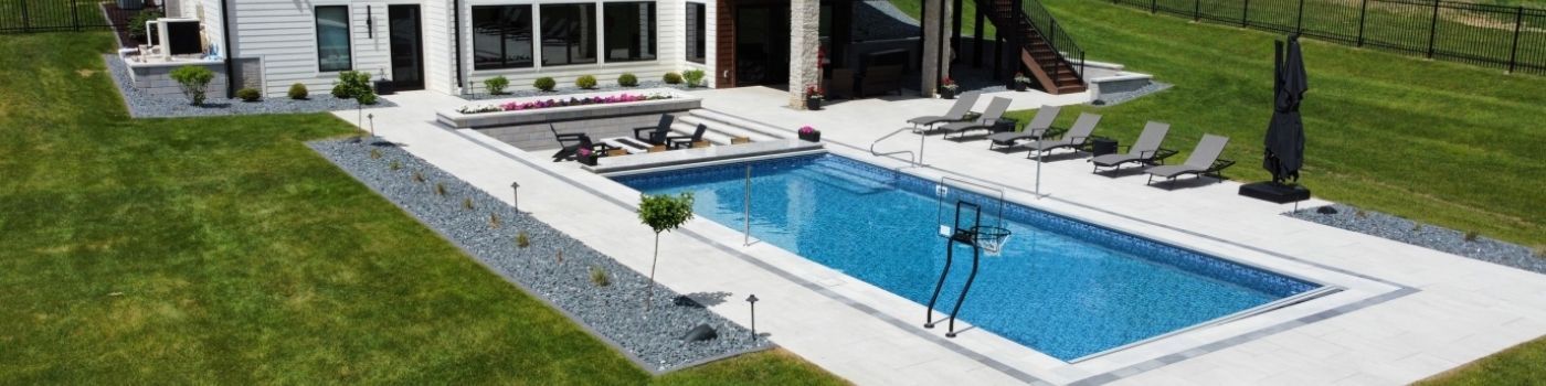 An aerial view of a rectangular pool surrounded by a white patio, lawn chairs, and lush green grass. A modern home is visible.