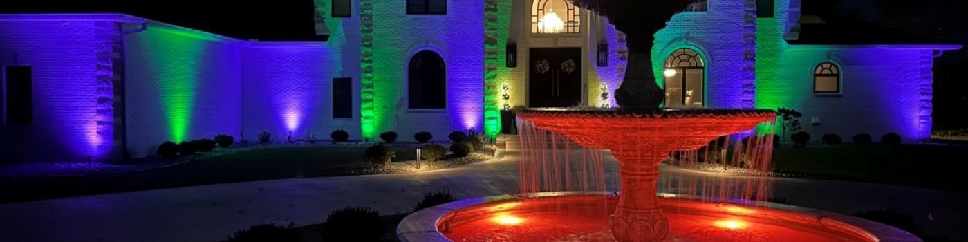 A nighttime view of a mansion with colored lights. A red fountain is in the foreground, and the building has purple and green illumination.