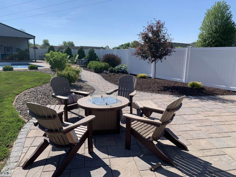 Four Adirondack chairs around a fire pit on a patio, with a pathway leading to a garden and a white fence.