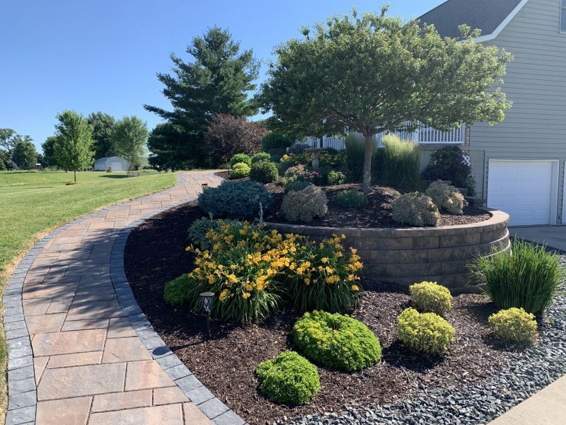 A stone path leads to a house with a landscaped garden. Yellow flowers and various green shrubs surround the curved flower bed.