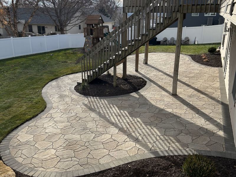 Stone patio with a curved walkway and stairs leading to a deck. The patio is next to a green lawn and a white fence.