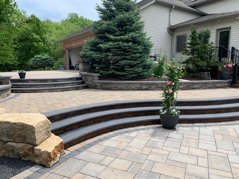 Outdoor patio with tiered steps, stone retaining walls, and a large evergreen tree near a house with a garage.