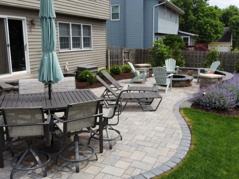 Backyard patio with a dining table, lounge chairs, and a fire pit, bordered by a brick pathway and landscaping.