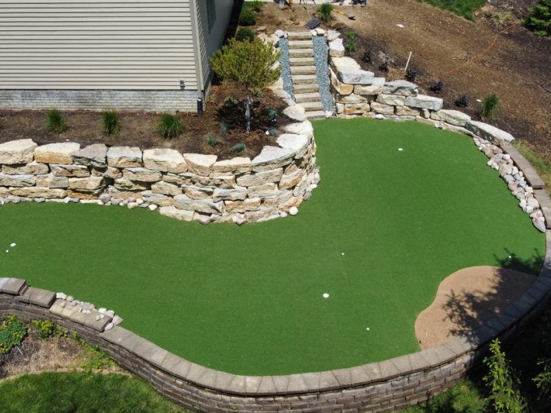 Artificial turf putting green with stone retaining walls and steps leading up to a house.