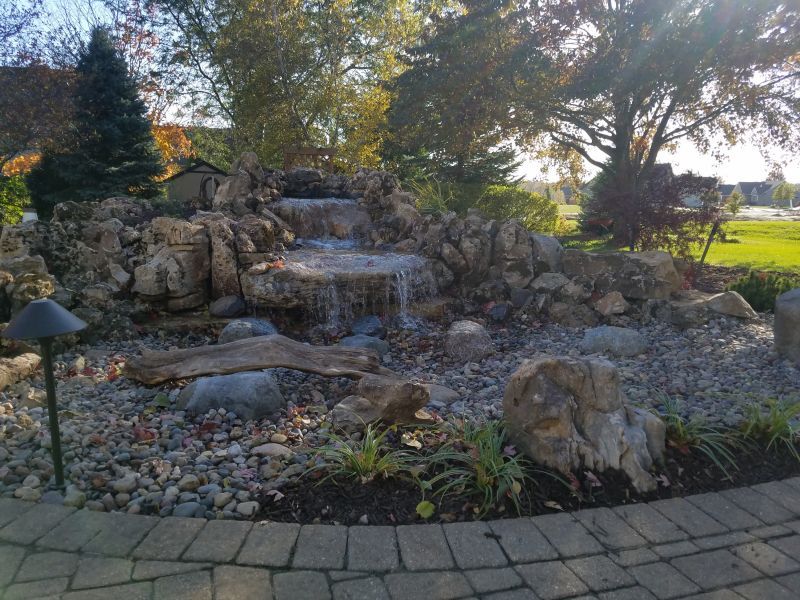 Water feature with cascading waterfalls built of rocks, surrounded by foliage. Located on a brick patio with greenery and trees in the background.