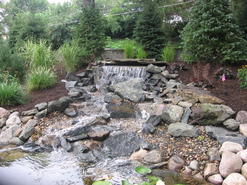A waterfall cascading over rocks into a pond, surrounded by plants and landscaping.