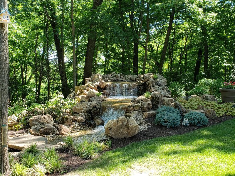 A tiered rock waterfall flows into a small pond, surrounded by greenery and trees.