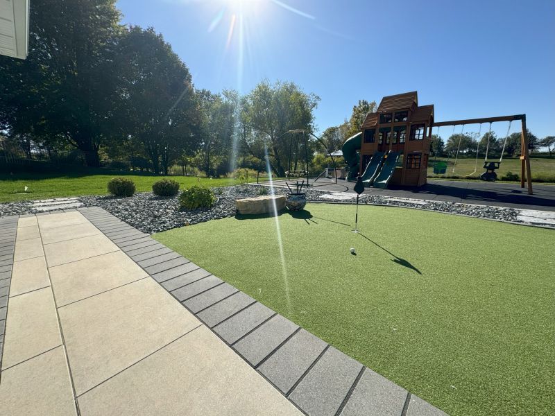 A sunny outdoor playground with a putting green in the foreground and a wooden play structure and swingset in the background.
