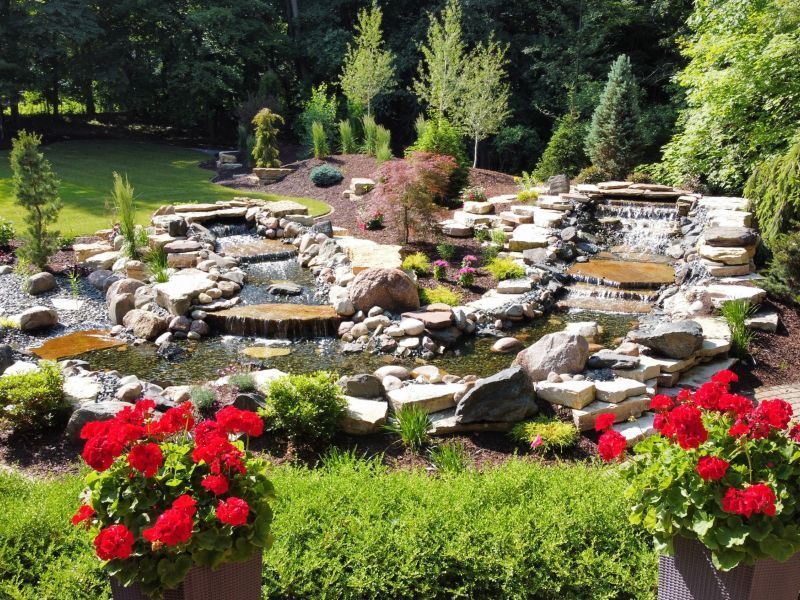 A tiered water garden with waterfalls, surrounded by rocks, greenery, and red flowers.